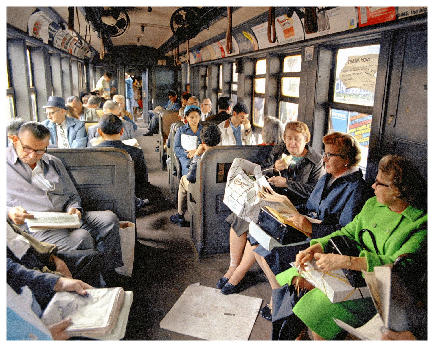 A vintage photograph of a crowded city bus interior with passengers seated in rows, reading newspapers and holding magazines. Passengers wear mid-century clothing, and posters and advertisements are visible along the bus walls and overhead.